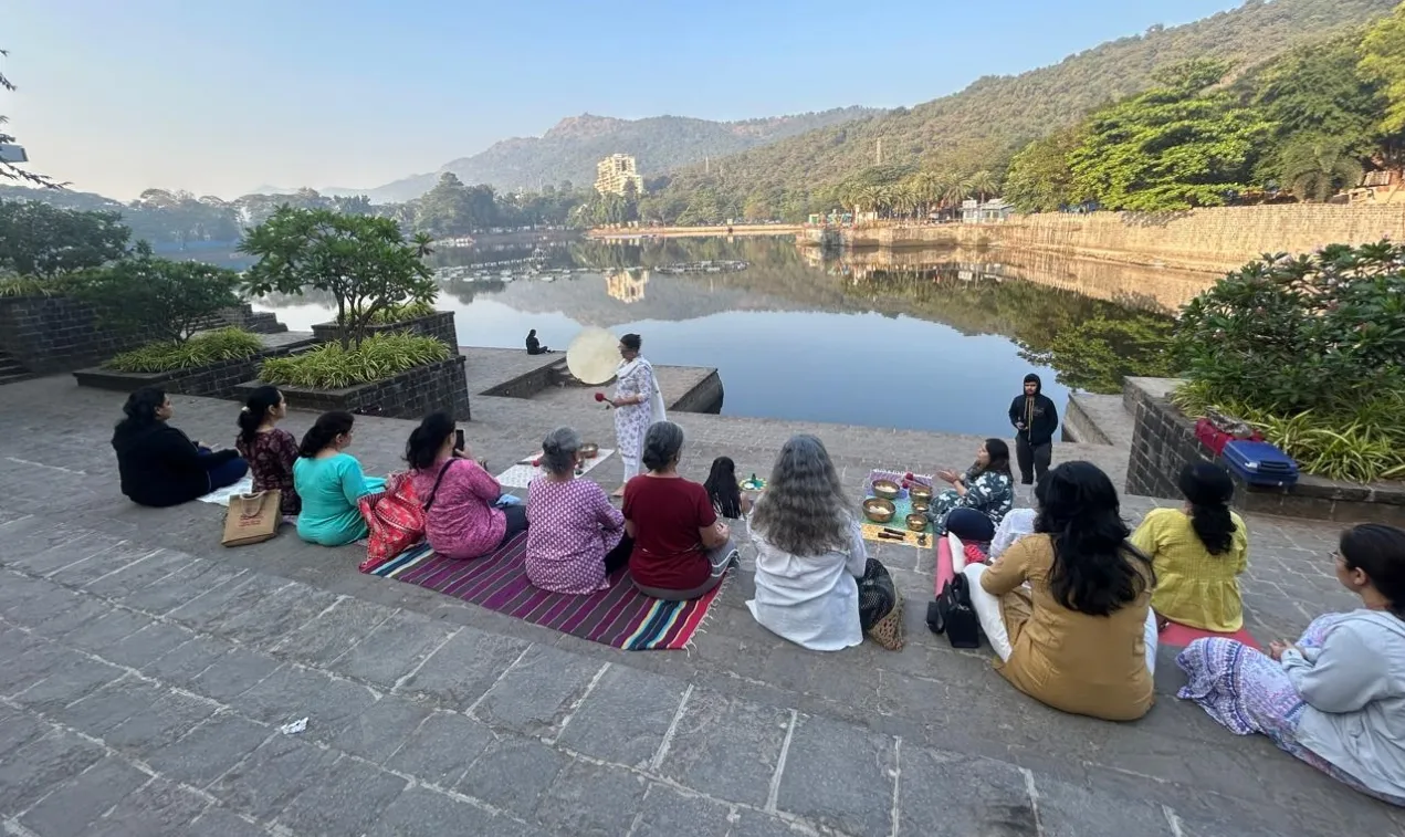 A group sound bath beside a lake in the morning, with participants seated on mats facing the water and hills